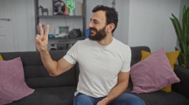 Neşeli orta yaşlı adam iki numarayı parmaklarıyla gösteriyor, beyaz tişört giyiyor, kendine güvenen bir gülümsemeyle yukarıyı işaret ediyor..