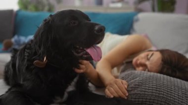 Oturma odasındaki kanepede evcil köpeği, siyah bir labrador ile dinlenen genç bir kadın, evde huzurlu bir anın tadını çıkarıyor..