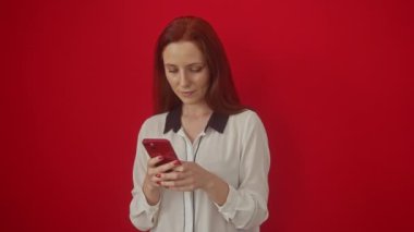 Şaşırmış genç beyaz kadın, ağzı açık, güvensizlik içinde, izole edilmiş kırmızı arka planda, akıllı telefon ekranında şaşkınlıkla bekliyor.