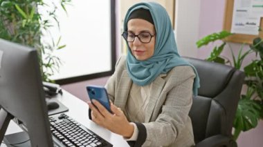 Başörtüsü takan profesyonel bir kadın modern ofis ortamında akıllı telefon kullanır, iş yerindeki çeşitliliği ve içeriği gösterir.