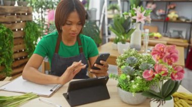 Afro-Amerikalı bir çiçekçi. Renkli bir çiçekçide akıllı telefon kullanıyor. Üzerinde yeşil önlük ve kapalı bitkiler var..