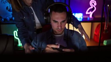 Geceleri neon ışıklı bir oyun odasında, bir kadın oturmuş, kulaklık takmış, video oyunu oynayan bir adamın arkasında gülümsüyor..