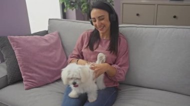 Kulaklık takan gülümseyen bir kadın oturma odasındaki kanepede bichon köpeğiyle vakit geçirir..