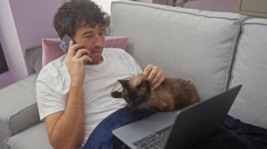 Genç bir adam telefonda konuşuyor. Kucağında kedi besliyor ve rahat bir oturma odasında dizüstü bilgisayar kullanıyor..