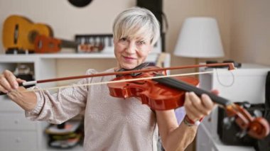 Gülümseyen kadın arka planda gitarlarla bir ev stüdyosunda keman çalıyor, neşe ve müzik tutkusu gösteriyor..