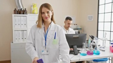 Elinde not panosu olan gülümseyen bir kadın bilim adamı laboratuvarda dikilirken erkek bir meslektaşı arka planda çalışıyor..