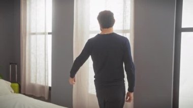 Orta yaşlı, sakallı bir adam modern bir yatak odasında neşeli bir ifadeyle durup perdeleri açar ve ışığı içeri alır..
