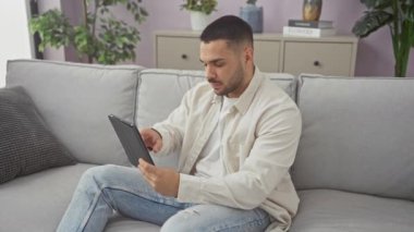 Evde gri bir kanepede rahatça otururken günlük kıyafetler giyen yakışıklı İspanyol bir adam tablet kullanıyordu.