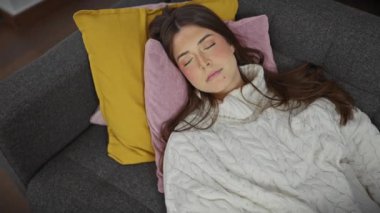 Huzurlu bir İspanyol kadın, gri bir kanepede rahat bir şekilde, renkli yastıklarla, kapalı bir odada dinleniyor.