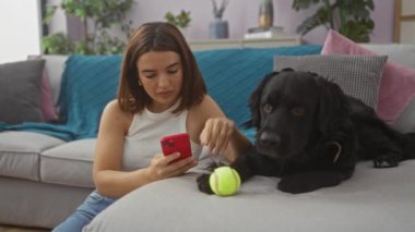 Genç bir İspanyol kadın akıllı telefon kullanıyor. Yanında siyah labrador köpeği ve tenis topu var. Oturma odasının rahat ortamında..