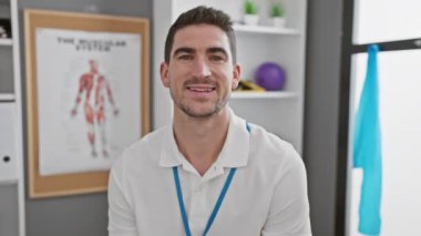 Beyaz polo tişörtlü, gülümseyen İspanyol bir adam arka planda kas sistemi çizelgesi olan bir terapi kliniğinde duruyor..