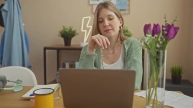 Düşünceli genç bir kadın, modern bir ev ofisinde dizüstü bilgisayarla bir masada oturur. Etrafı bitkiler ve sıradan dekorlarla çevrilidir..