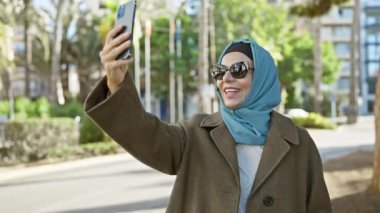 Orta yaşlı tesettürlü gülümseyen bir kadın akıllı telefonuyla bir avuç palmiye ağacına ve şehir sokaklarına selfie çekiyor..