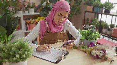 Pembe tesettürlü bir kadın panoya yazı yazar ve bitkilerle dolu bir odada akıllı telefon kullanır..