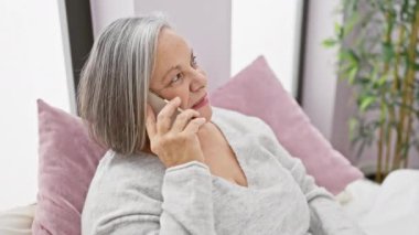 Üst düzey kadın akıllı telefonda konuşuyor, yatağın içinde dinleniyor, yaşlılar tarafından konfor ve teknoloji kullanımı sergiliyor.