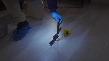 Koruyucu ekipman adli tıp uzmanı loş ışıklı bir suç mahallinde zeminde numaralandırılmış bir işaretle kanıtları inceliyor..