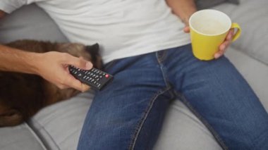 Elinde kahve fincanı ve uzaktan kumandası olan bir adam koltukta uyuyan siyam kedisinin yanında gevşiyor..