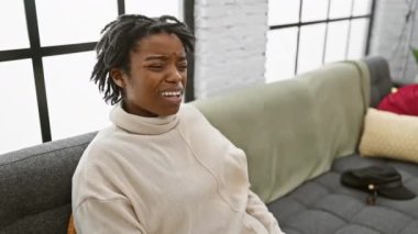 Rasta saçı olan ürkmüş genç siyahi bir kadın rahat bir ev kanepesinde oturmuş, şaşkın bakışlarıyla, alaycı bir inançsızlıkla ağzı sarkık..