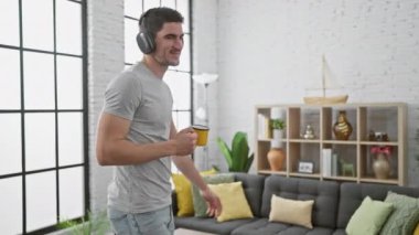 Yakışıklı genç bir adam evinde elinde bir kupayla, kulaklıkla şık, modern bir oturma odasında neşeyle dans ediyor..