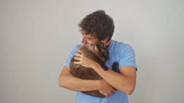 Genç bir adam beyaz bir arka planda sevgi dolu bir siyam kedisini kucaklıyor.