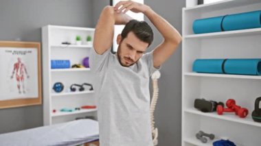 Genç sakallı bir adam rehabilitasyon kliniğinde spor malzemeleriyle dolu bir odada geriniyor..