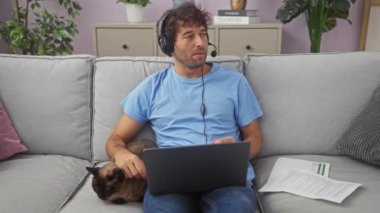 Kulaklıklı düşünceli bir adam rahat bir oturma odasında kanepede kedi beslerken dizüstü bilgisayarda çalışıyor..
