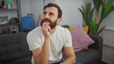 Düşünceli orta yaşlı bir adam, beyaz bir tişört giyiyor, elleri çenesinde, evinde derin bir düşünme seansına dalmış, şüphe denizinde yol alırken yüzünde düşünceli bir ifade var.