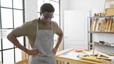 Güvenlik gözlüklü, kendine güvenen genç bir siyah adam, iyi ışıklandırılmış marangozluk atölyesinde duruyor..