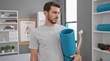 Neşeli İspanyol genç adam dişlek gülümseme saçıyor, rehabilitasyon kliniğinde yoga minderi tutuyor.