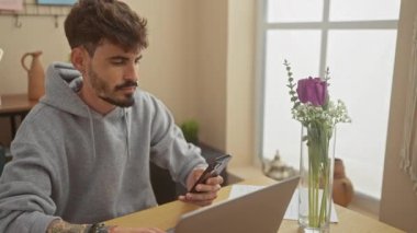 Hoş bir dairede akıllı telefon ve dizüstü bilgisayar kullanan dövmeli yakışıklı bir İspanyol..