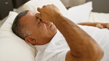 Beyaz tişörtlü, orta yaşlı bir İspanyol, bir yatakta yatar. Alnını tutarken acı ya da rahatsızlık belirtileri gösterir..