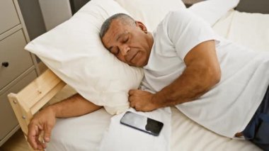 Beyaz gömlekli orta yaşlı İspanyol bir adam yatak odasında yanında bir telefonla huzur içinde uyuyor..