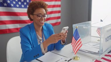 Mavi ceketli Afro-Amerikalı bir kadın akıllı telefon kullanırken gülümsüyor. Oy sandığının yanında oturuyordu..