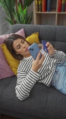 Genç İspanyol kadın akıllı telefon kullanıyor ve içeride koltukta yatarken elinde kredi kartı tutuyor.
