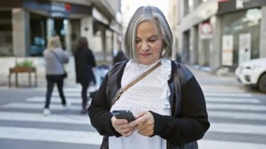 Şehir caddesinde akıllı telefon kullanan, konsantrasyonunu ve günlük tarzını ifade eden kıdemli bir kadın..