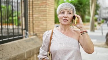 Gri saçlı olgun bir kadın açık hava şehrinde telefonla konuşuyor..