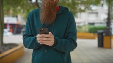 Akıllı telefonlu sakallı bir adam şehir parkında dikilip şehir yaşamını gözler önüne seriyor..