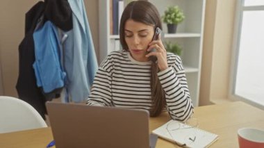 Odaklanmış genç bir İspanyol kadın telefonda konuşurken dizüstü bilgisayarıyla çalışıyor. Çoklu görev ve uzak çalışma yaşam tarzını somutlaştırıyor.