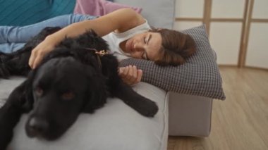 Genç bir İspanyol kadın oturma odasındaki kanepede huzur içinde uyuyor, siyah labrador köpeğine sarılıyor..