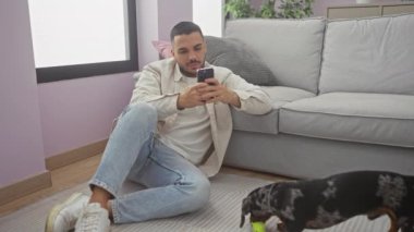 Günlük giysiler içinde genç bir İspanyol adam evde akıllı telefon kullanıyor ve yanında da şakacı bir daksund 'u var..