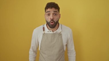 Şaşırmış, ağzı açık, inanamamaktan korkan, sarı arka planda önlük takan genç İspanyol bir adam, yüzündeki şaşırmış ifadeyi açığa vuruyor.