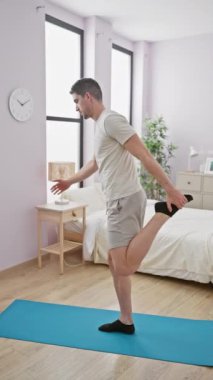 Parlak bir yatak odasında yoga yapan bir adam mavi bir paspasın üzerinde odak ve esneklik ile dengeyi koruyor..