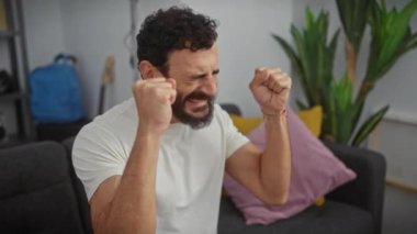 Neşeli orta yaşlı bir adam, galibiyete sevinmiş, evinde zaferden zevk alıyor, beyaz bir tişörtle, geniş bir gülümseme ve gözleri kapalı, neşeli kollar kaldırılmış.