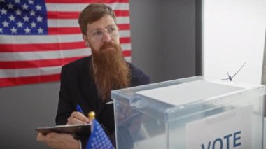 Gözlüklü sakallı bir adam, kapalı alanda Amerikan bayrağına karşı oy sandığına bakıyor..