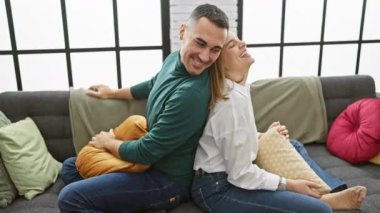 Gülümseyen bir erkek ve kadın, büyük pencereli, güneşli bir oturma odasındaki kanepede neşeyle kucaklaşır..