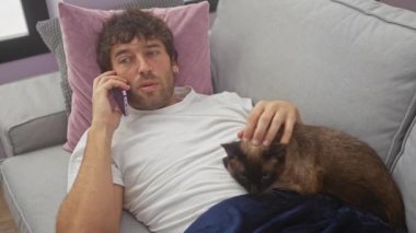 Sıcak bir oturma odasında kedisini okşarken telefonda konuşan stresli, genç İspanyol bir adam..