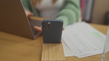 Gözlüklü sarışın bir kadın akıllı telefonu ve laptopuyla evdeki belgeleri inceliyor, odaklanmış iş ve teknoloji kullanımını çağrıştırıyor.