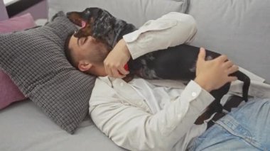 Günlük giysiler içinde genç bir İspanyol adam evde bir kanepede dachshund 'uyla eğleniyor..