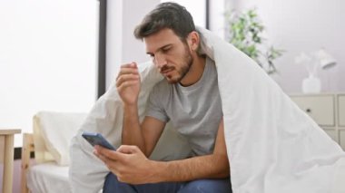 Endişeli genç bir İspanyol, yatak odasındaki akıllı telefonuna bakarken bir yatakta başını tutarak oturur..