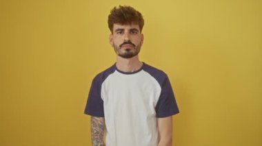 İzole edilmiş sarı bir arka planda duran kendine güvenen genç bir İspanyol, kameraya gülümseyerek bakıyor, kolları çapraz ve elleri çenede, pozitif düşünceyi yansıtıyor.